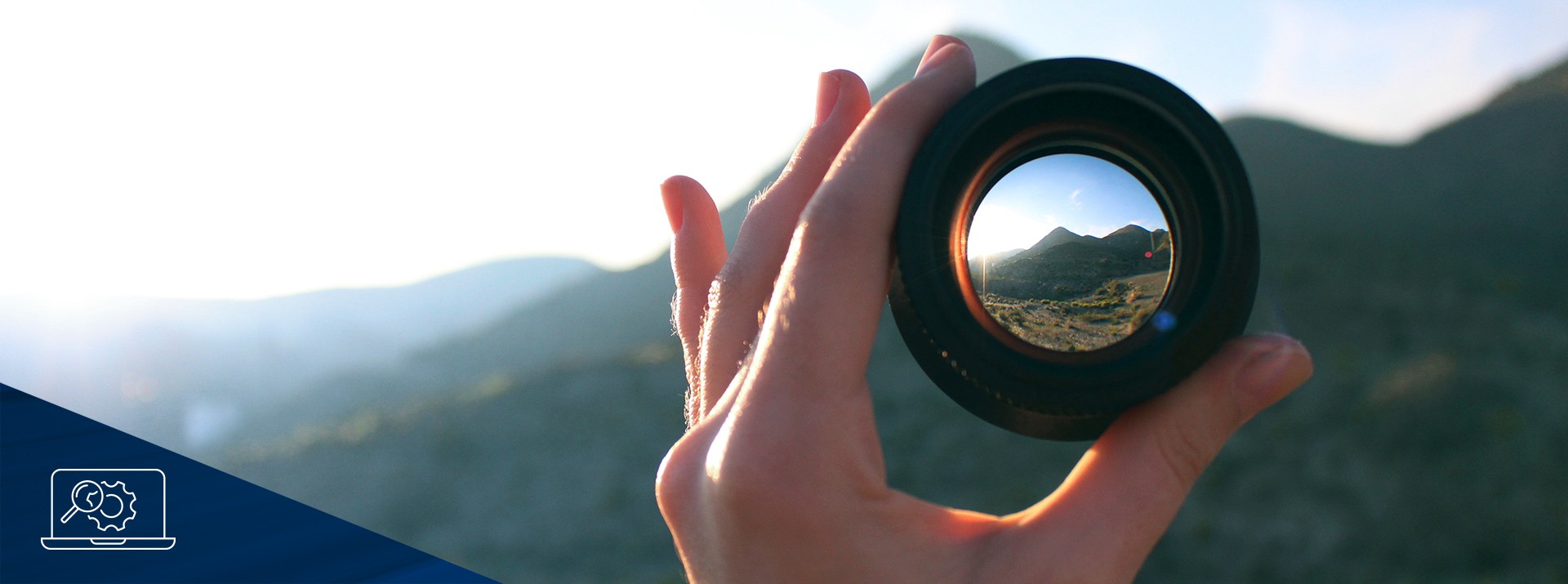 Person holding camera lens towards mountain landscape.