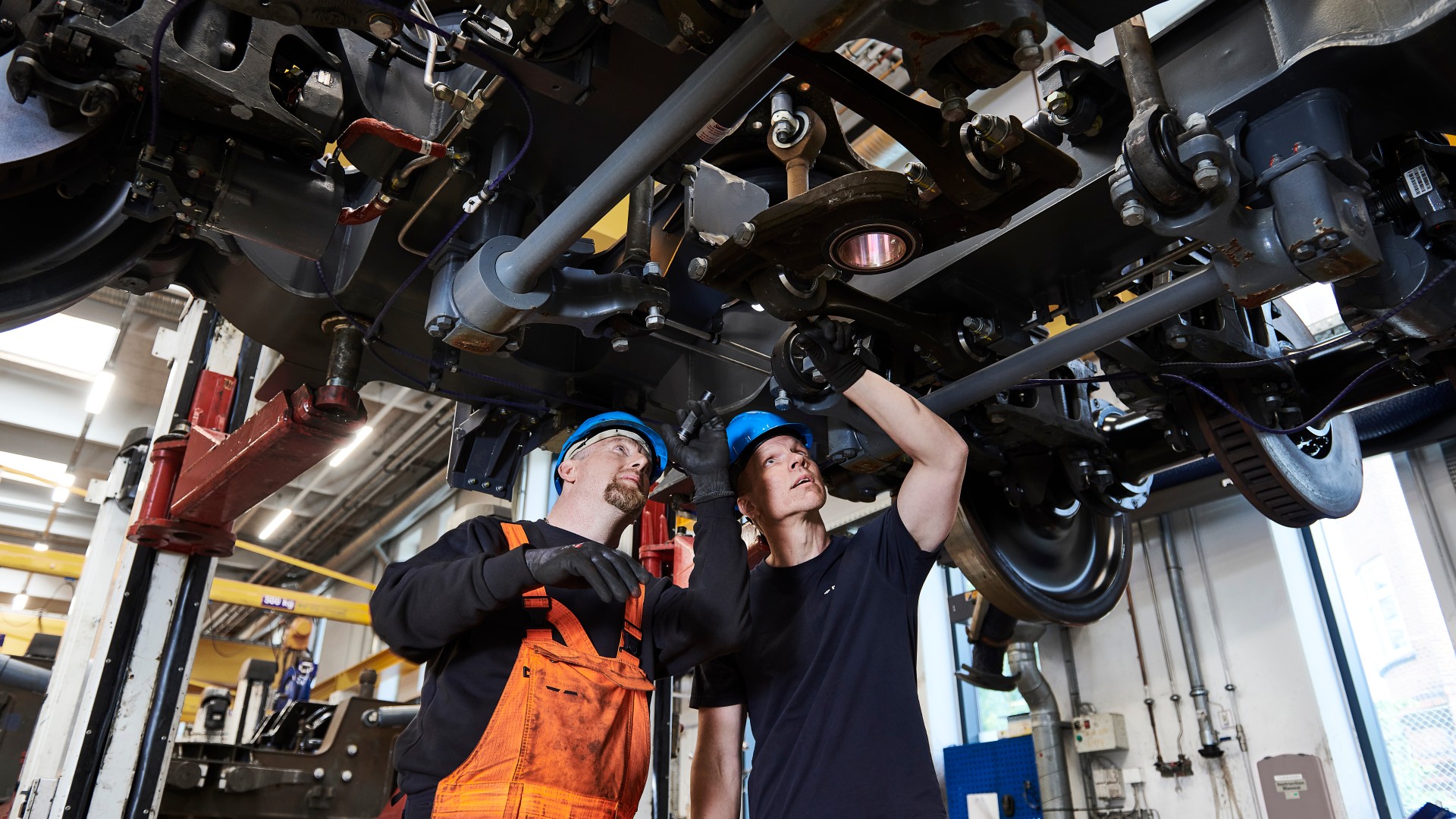 Two men under a train looking at the mechanical components