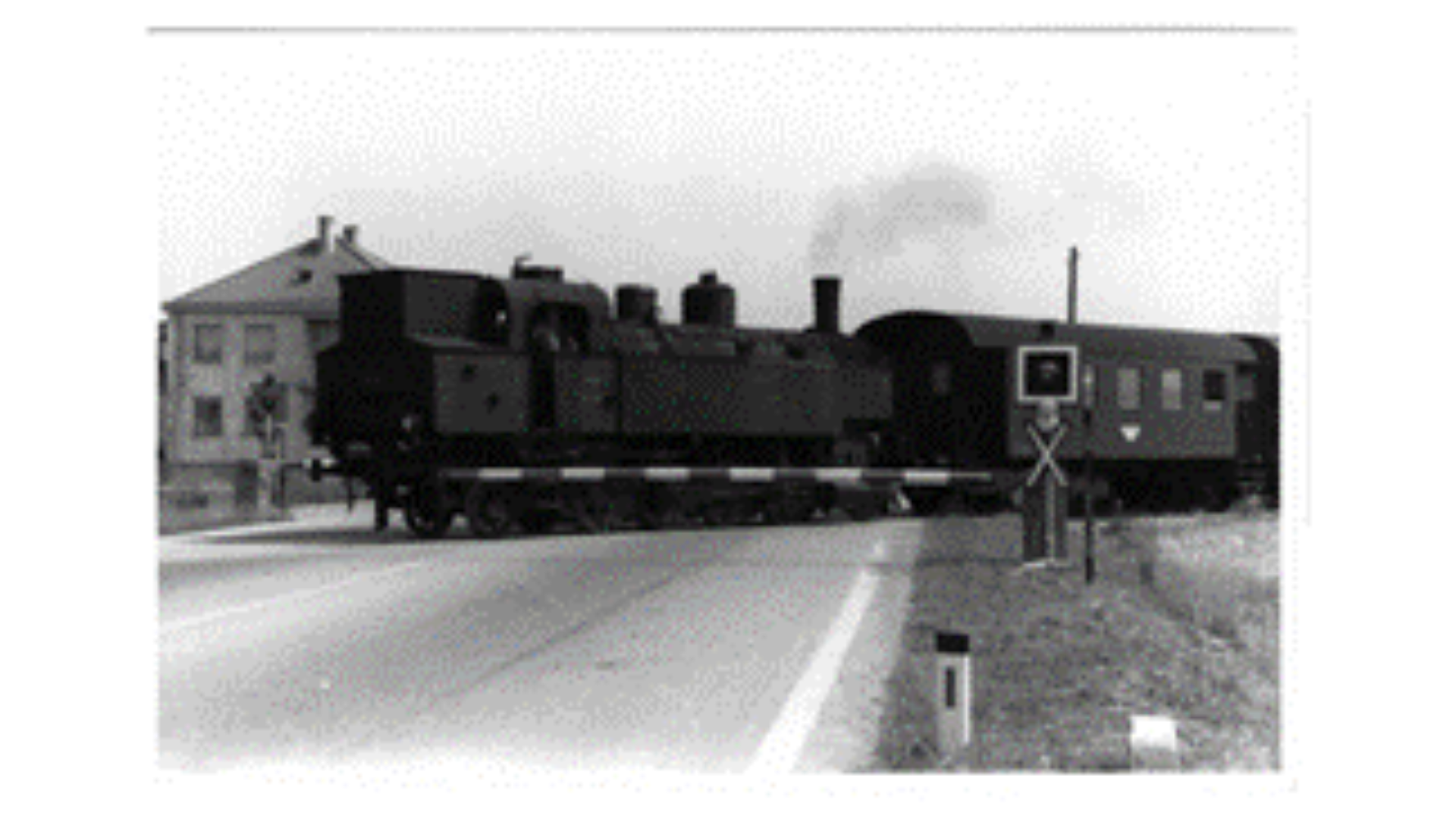 Steam locomotive crossing a road near buildings.