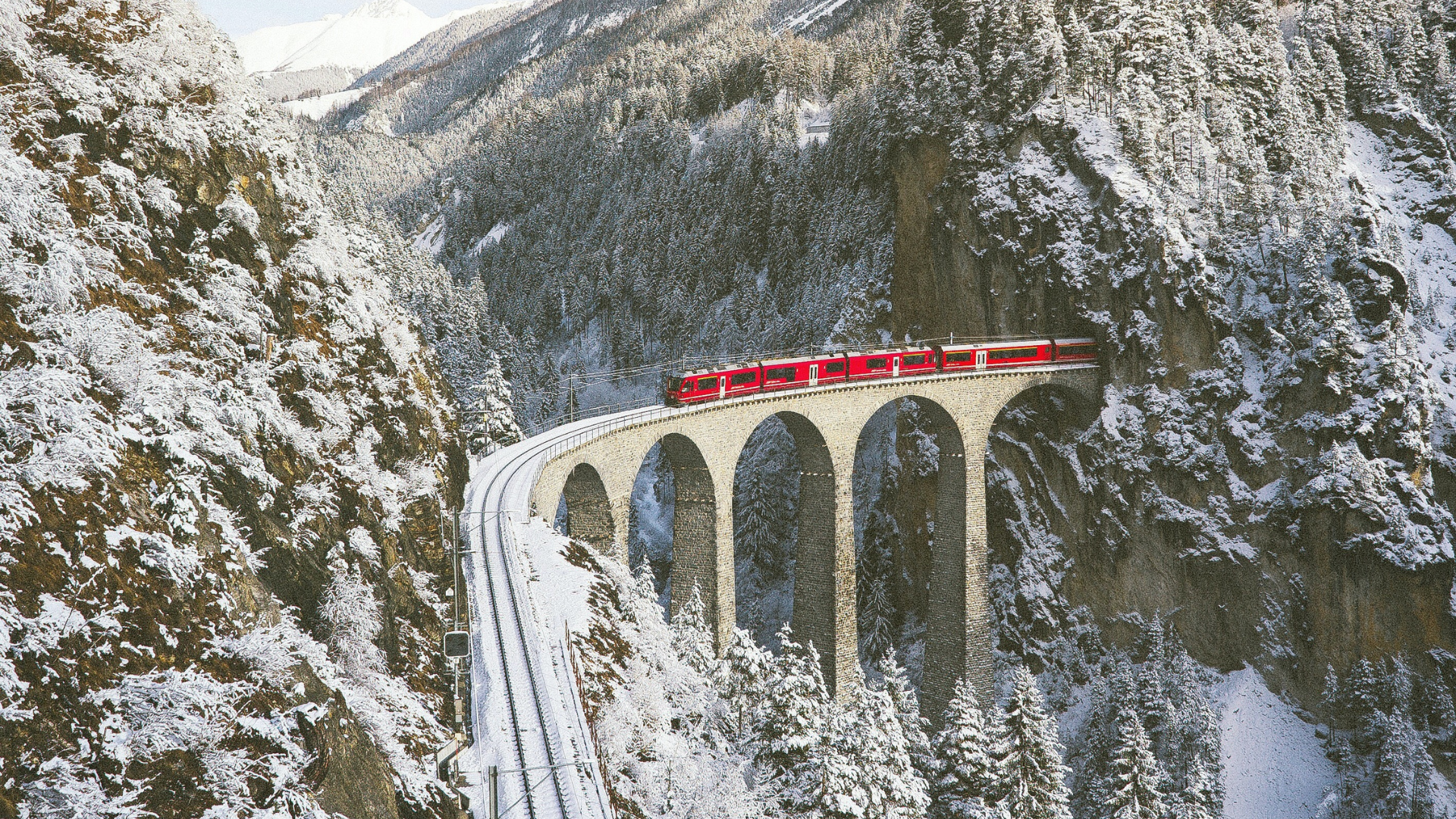 Red train on snow-covered mountain viaduct.